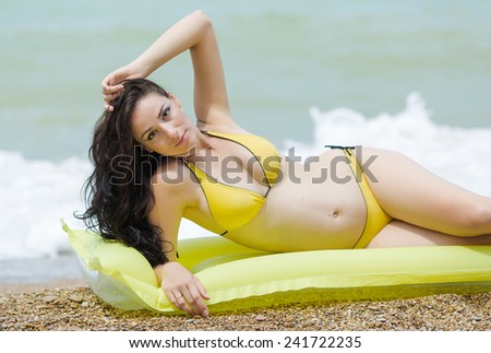 Expectant mother lying on pool raft looking at camera. Pregnant woman in yellow bikini on pebble beach