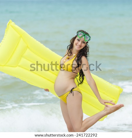Expectant mother with pool raft and diving mask looking at camera. Pregnant woman in yellow bikini playing on the beach