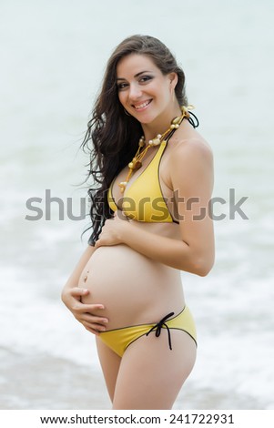 Expectant mother embracing own tummy looking at camera smiling. Pregnant woman in yellow bikini at the sea