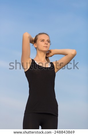 Young woman in black sportswear stands with arms raised. Slim girl corrects own hairstyle looking away