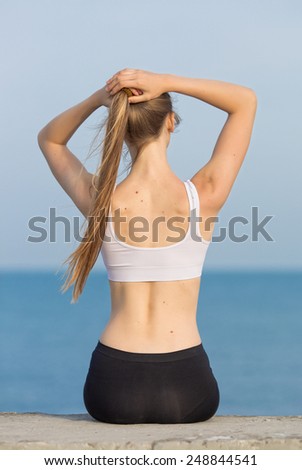 Young woman in sportswear at the sea. Slim girl sitting on concrete pier corrects own ponytail, rear view