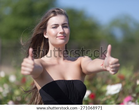 Waist up portrait of girl showing two thumbs up looking at camera. Young long-haired woman in black sleeveless dress expressing positive emotions outdoors