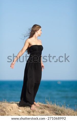 Barefoot young woman in black at the sea. Long haired girl in black long dress stands on seashore looking away