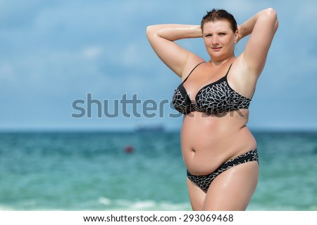 Woman in swimwear at the sea. Overweight young woman in swimsuit posing against horizon over water. Female with arms raised corrects her hair looking at camera
