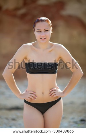 Girl on the beach. Young woman in black swimwear posing arms akimbo on on seashore