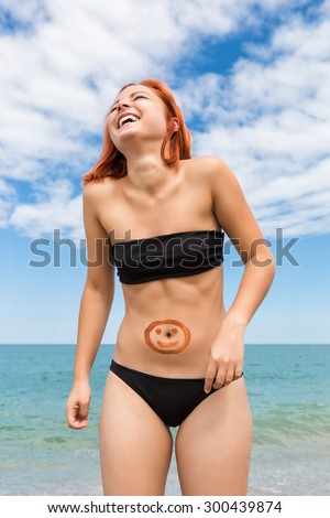 Young woman at the sea. Laughing ginger girl with a painted smiley on her belly against the sea