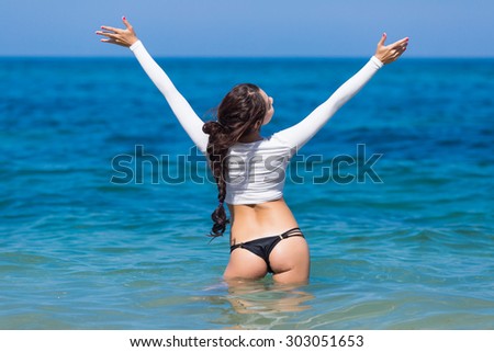 Girl at the sea. Slim brunette in white crop top with long sleeves stands in water with arms raised looking up