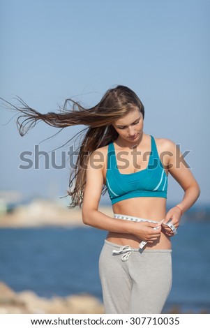 Girl measures her waist looking down. Attractive young woman in sports bra with tape-measure on background of sky