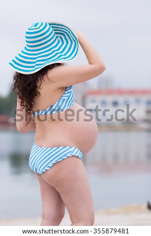 Expectant mother on seashore. Pregnant woman in striped bikini and striped hat stands on beach looking away