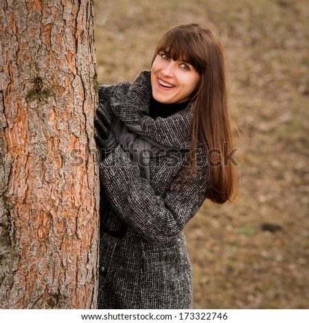 Beautiful woman in stylish winter fashion wearing a long belted knitted jumper and gloves hiding behind of a pine tree outdoors and smiling at the camera