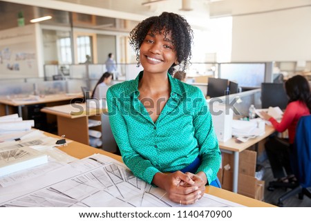 Black female architect leans on desk smiling in busy office