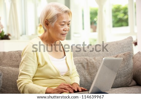 Senior Chinese Woman Using Laptop Whilst Relaxing On Sofa At Home