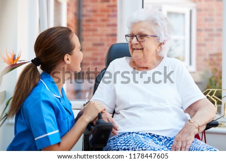 Senior Woman Sitting In Motorized Wheelchair Talking With Nurse In Retirement Home