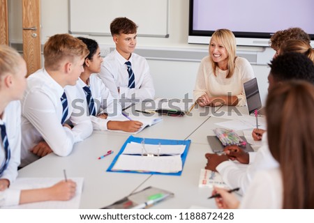Female High School Teacher Sitting At Table With Teenage Pupils Wearing Uniform Teaching Lesson