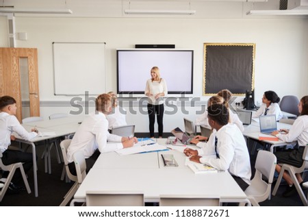 Female High School Teacher Standing Next To Interactive Whiteboard And Teaching Lesson To Pupils Wearing Uniform