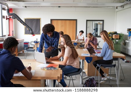 Teacher Talking To Female High School Student Sitting At Work Bench Using Laptop In Design And Technology Lesson