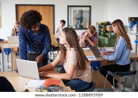 Teacher Talking To Female High School Student Sitting At Work Bench Using Laptop In Design And Technology Lesson