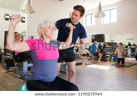 Senior Woman Exercising On Swiss Ball With Weights Being Encouraged By Personal Trainer In Gym