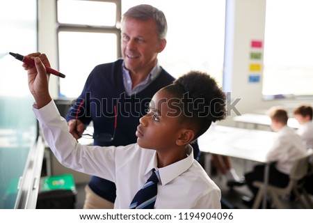 Male High School Teacher With Female Student Wearing Uniform Using Interactive Whiteboard During Lesson