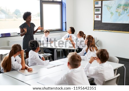 Female High School Tutor Standing By Tables With Students Wearing Uniform Teaching Lesson