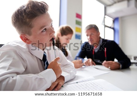 Concentrating Male High School Student Wearing Uniform Working At Table With Teacher Talking To Pupils In Background