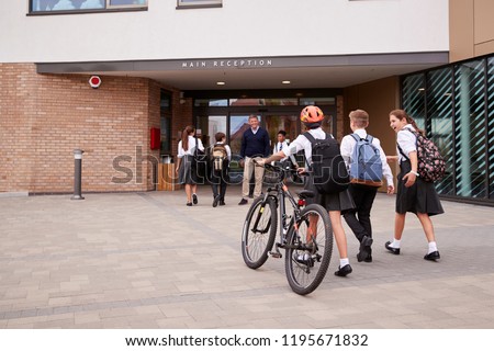 Group Of High School Students Wearing Uniform Arriving At School Walking Or Riding Bikes Being Greeted By Teacher