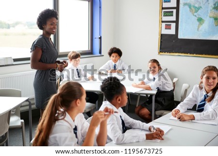 Female High School Tutor Helping Students Wearing Uniform Seated Around Tables In Lesson