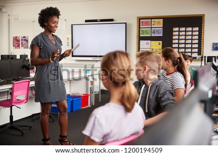 Teacher With Digital Tablet Talking To Line Of High School Students Sitting By Screens In Computer Class