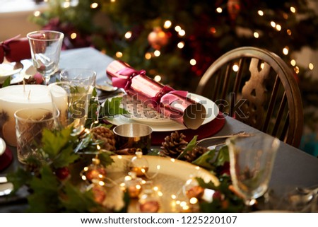 Christmas table setting with a Christmas cracker arranged on a plate with red and green table decorations and a Christmas tree in the background