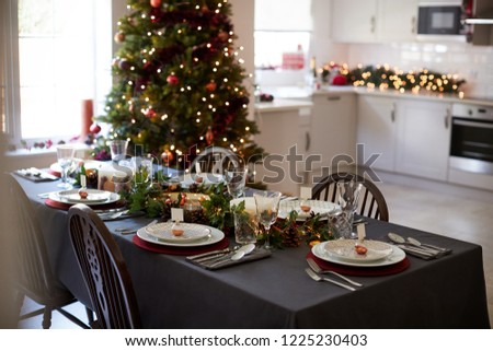 Christmas table setting with bauble name card holders arranged on plates and green and red decorations with Christmas tree and kitchen in the background