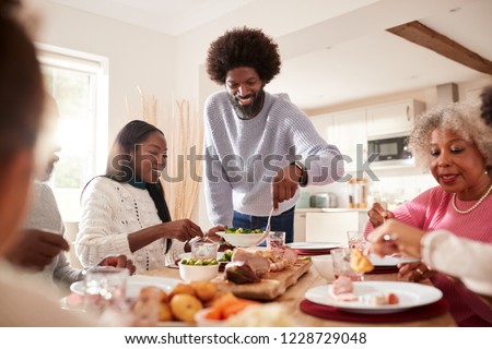 Middle aged black man carving and serving meat at Sunday family dinner with his partner, kids and their grandparents, front view