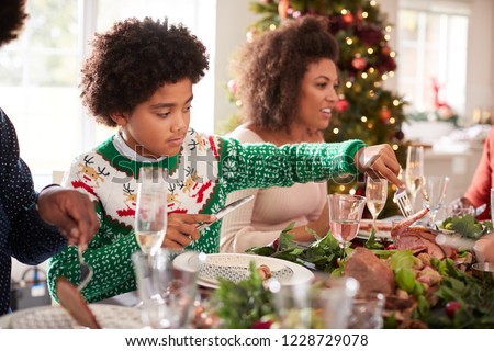 Tween black boy serving himself roast turkey sitting at the Christmas dinner table with his family, side view