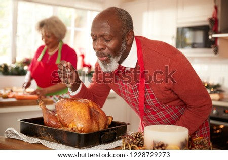 Senior black man basting roast turkey in preparation for Christmas dinner, his wife chopping vegetables in the background, close up, selective focus