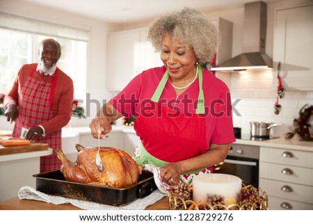 Senior black woman basting a roast turkey in preparation for Christmas dinner, her husband chopping vegetables in the background, front view, close up