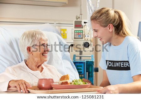 Teenage Volunteer Serving Senior Female Patient Meal In Hospital Bed