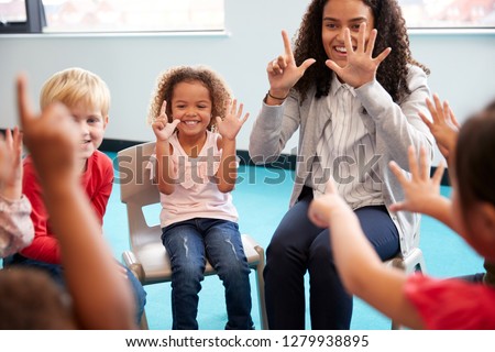 Front view of infant school children sitting on chairs in a circle in the classroom, holding up their hands and learning to count with their female teacher, close up