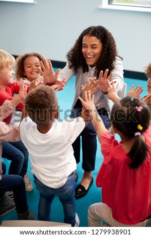 Elevated view of infant school children in a circle in the classroom giving high fives to their smiling female teacher, vertical, close up