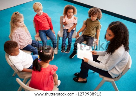 Young female school teacher reading a book to infant school children, sitting on chairs in a circle in the classroom listening, elevated view