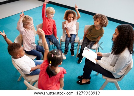 Young female school teacher reading a book to infant school children, sitting on chairs in a circle in the classroom raising hands to answer a question, elevated view