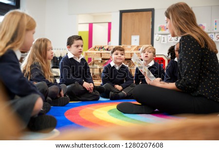 Female primary school teacher sitting on the floor in a classroom reading a book to her class, selective focus