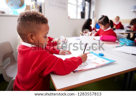 Young black schoolboy wearing school uniform sitting at a desk in an infant school classroom drawing, close up, side view