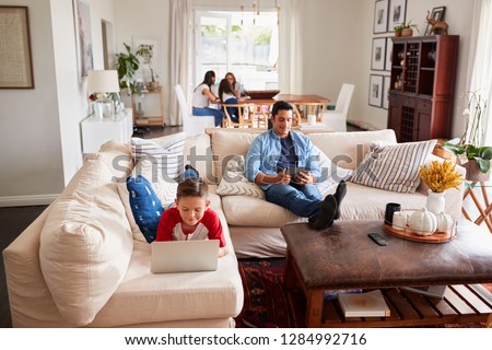 Pre-teen boy lying on sofa using laptop, dad using tablet, mum, sister and grandma in the background