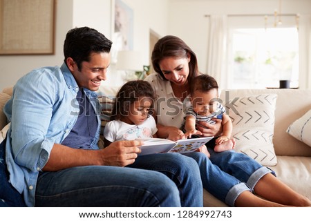 Young Hispanic family of four sitting on the sofa reading a book together in their living room
