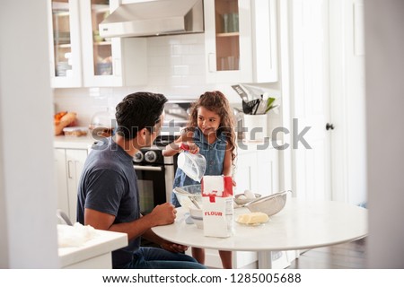 Young girl standing at the kitchen table preparing cake mixture with her father, seen from doorway