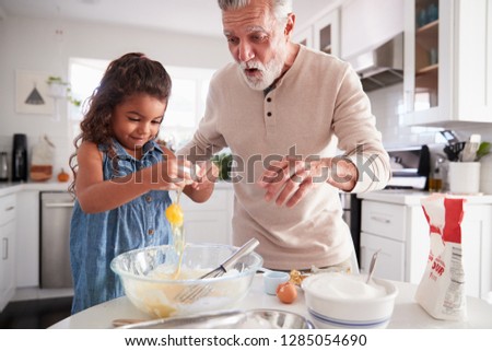 Young girl breaking an egg into cake mixture with her grandfather at the kitchen table, close up