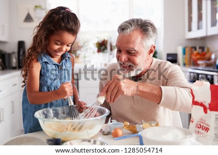 Young Hispanic girl and her grandad whisking cake mixture together at the kitchen table, close up