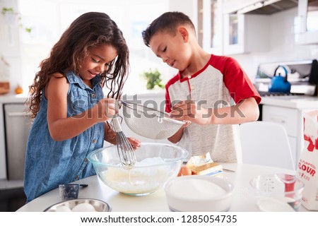 Brother and sister preparing cake mixture together at the kitchen table, waist up