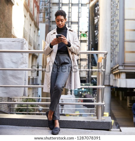 Fashionable young black woman standing in the city leaning on a hand rail using her smartphone, full length, low angle