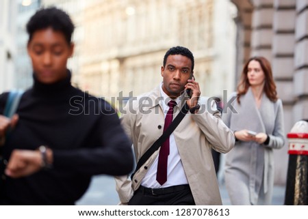 Millennial black businessman walking in a busy London street using smartphone, selective focus