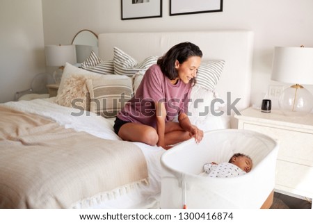 Young adult African American mother sitting on her bed looking down at her three month old baby sleeping in his cot